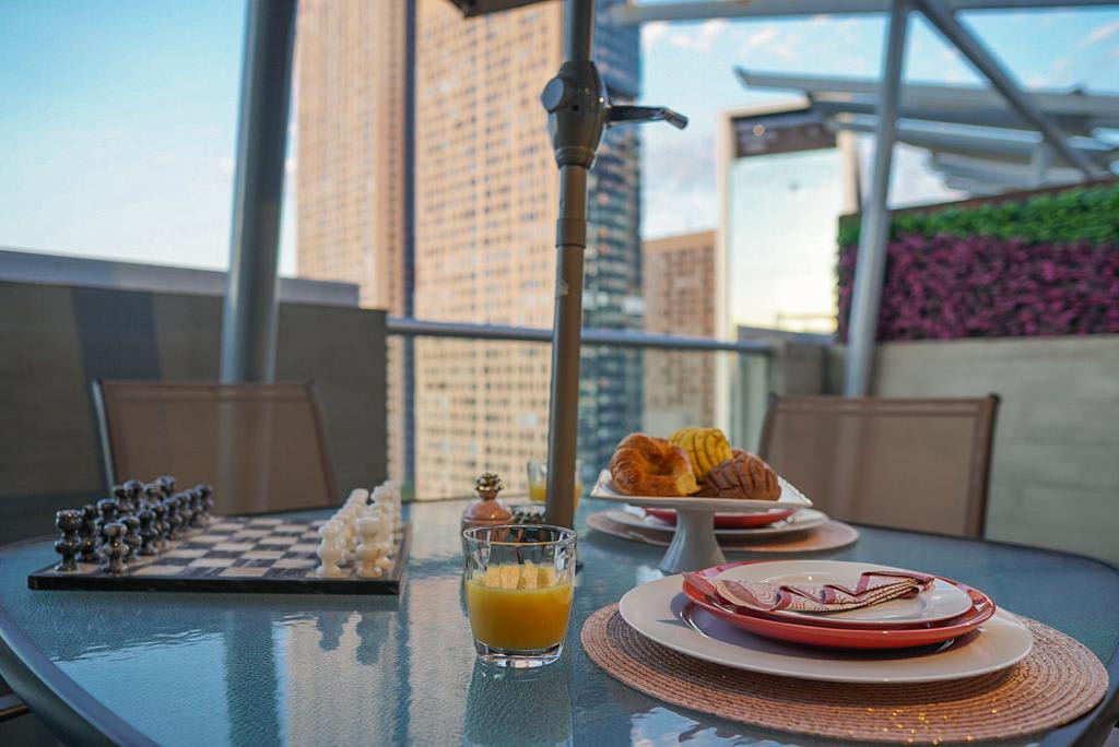 A dining table is set on a rooftop terrace, featuring a glass surface with colorful plates, a small glass of orange juice, and a platter of pastries. A chessboard is positioned nearby against a backdrop of modern high-rise buildings and greenery.
