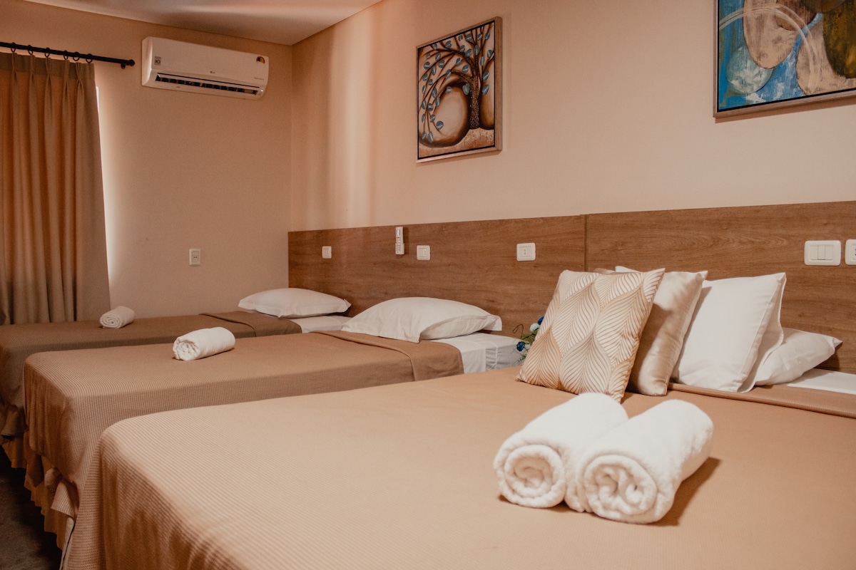 A spacious room features three neatly made beds adorned with neutral-toned linens. Soft towels are rolled and placed at the foot of each bed. Simple artwork decorates the walls, complemented by warm lighting from the overhead fixtures.