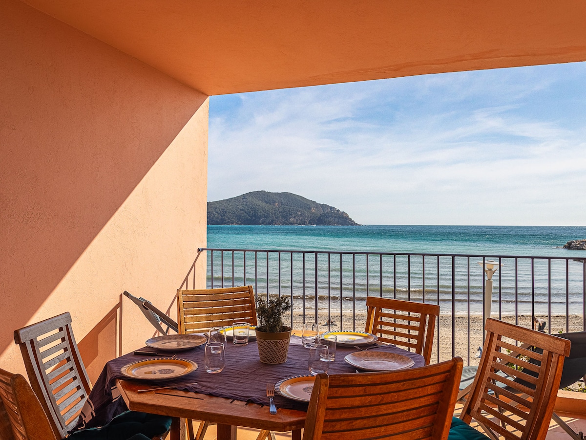 A large balcony features wooden dining furniture, set with tableware. The ocean view is visible beyond the railing, showcasing sandy beaches and distant hills. Natural light brightens the space, highlighting the inviting atmosphere.
