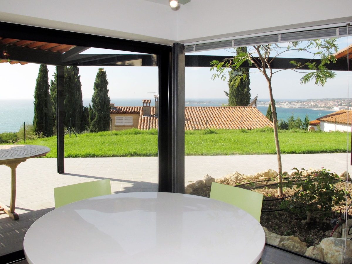 A modern dining area is presented with a round white table and light green chairs. Large glass panels provide a panoramic view of the sea and surrounding landscape. The space is illuminated by natural light, enhancing the connection to the outdoor scenery.