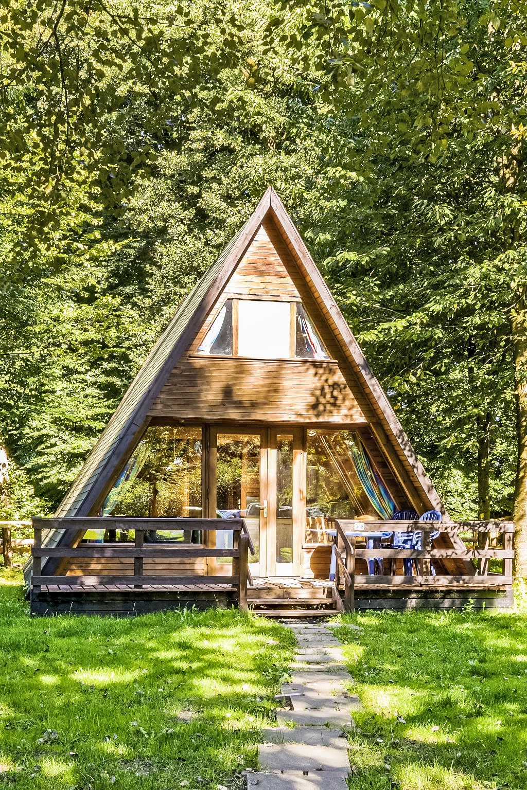 The A-frame house is surrounded by trees, showcasing a wooden exterior with large windows. A shaded terrace is visible, furnished with tables and chairs. A stone pathway leads up to the entrance, enhancing access to this nature-embedded retreat.