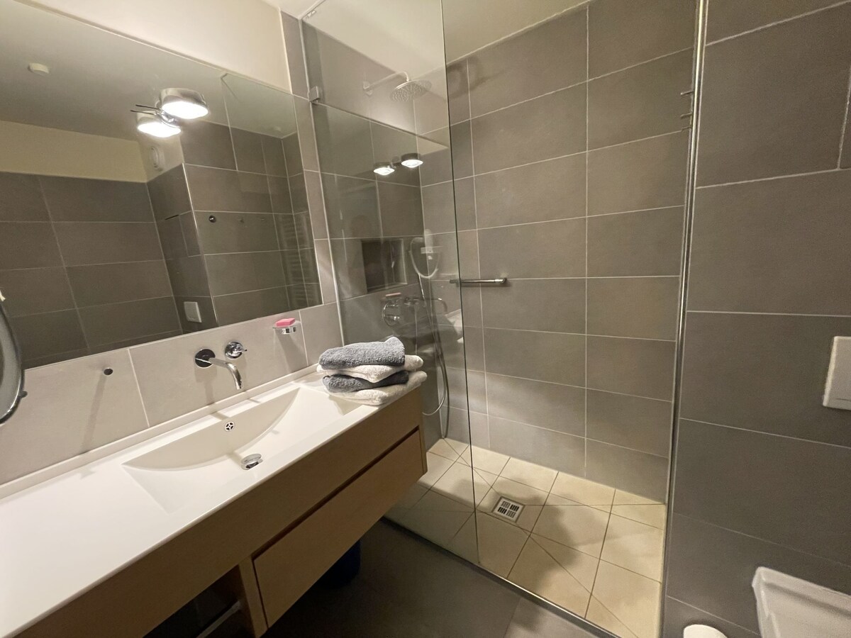 A modern bathroom features a walk-in shower with clear glass doors and neutral-toned tiles. A white vanity with a sink is positioned against the wall, complemented by a large mirror and two light fixtures above. Fresh towels are neatly arranged on the countertop.