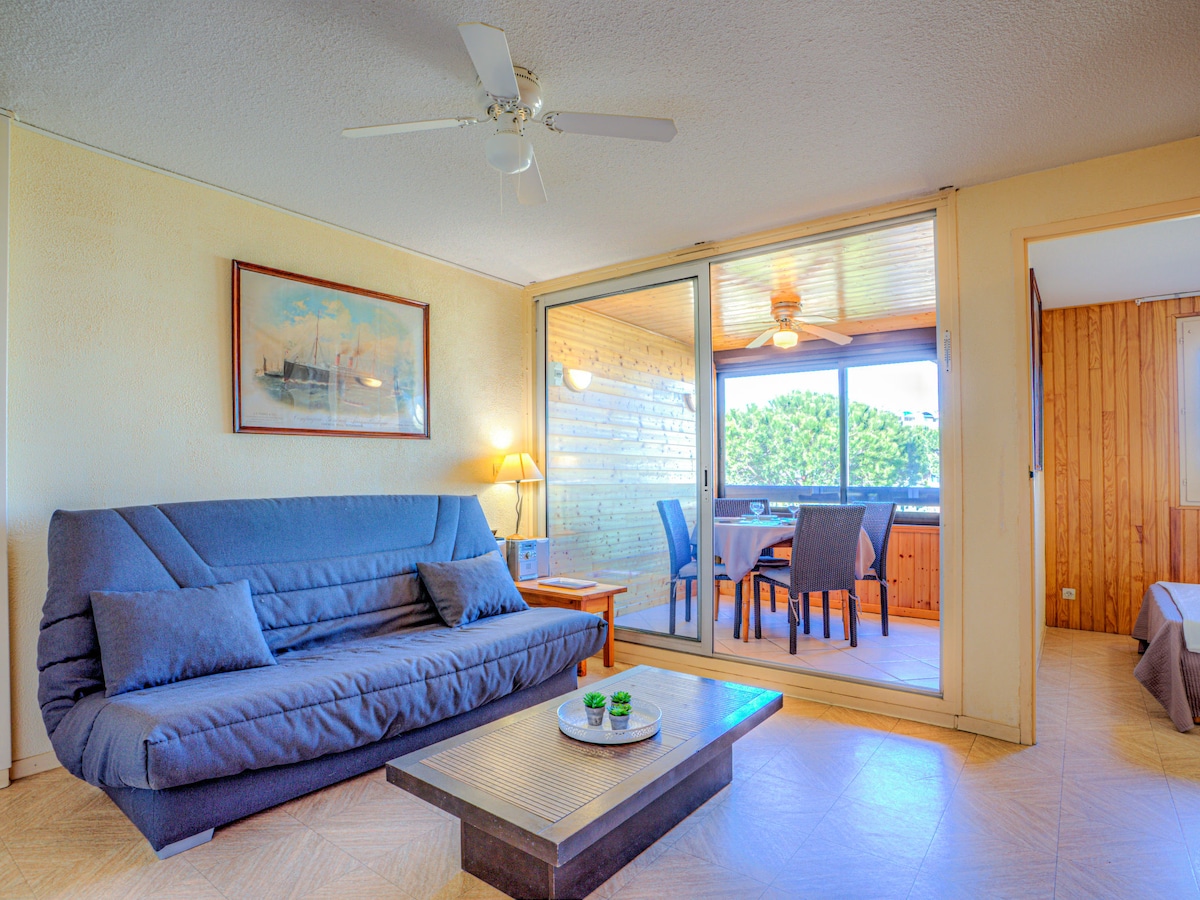 A living room area is shown with a blue sofa bed facing a wooden coffee table. A lamp provides soft lighting next to the window. Glass doors lead to an enclosed balcony with a dining table and chairs, overlooking natural greenery.