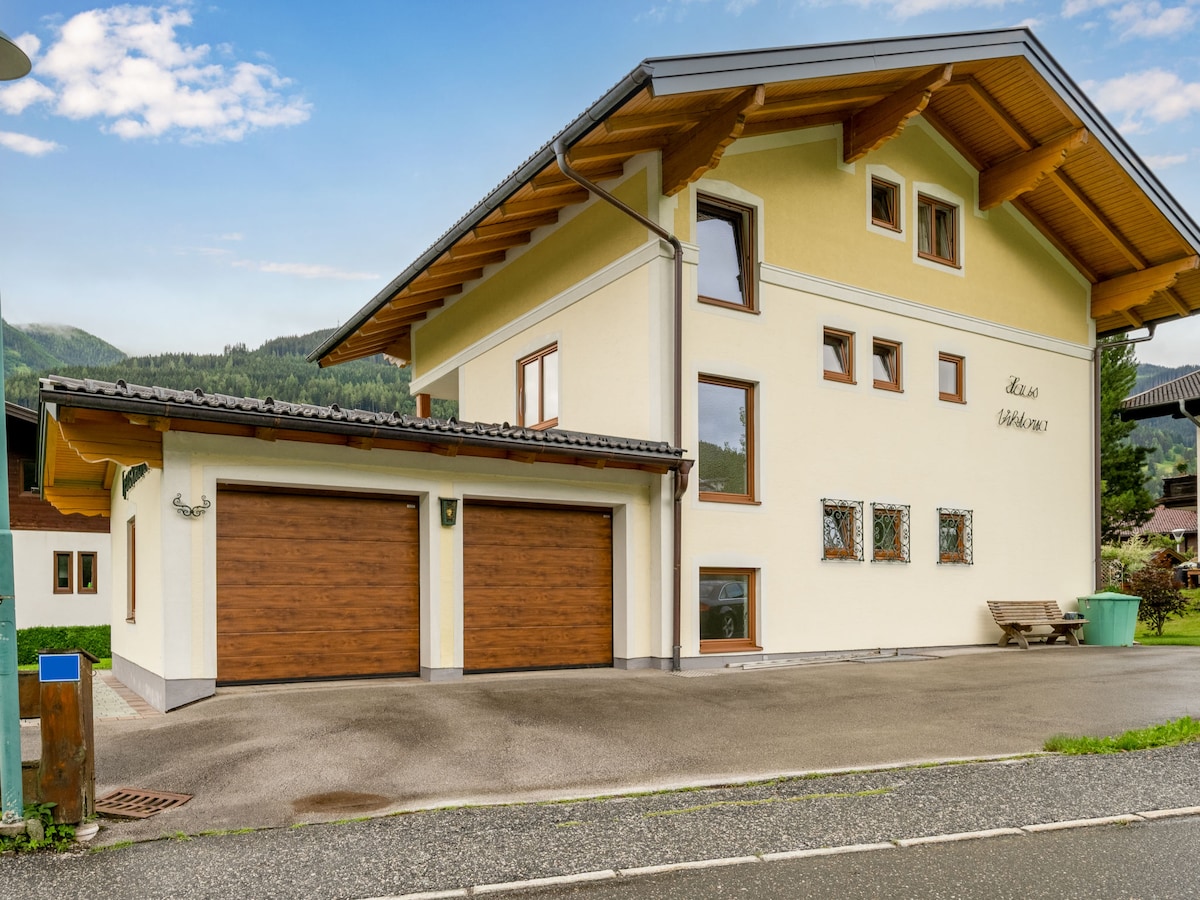 The exterior of the two-family house features a warm yellow facade, complemented by a sloping wooden roof. Two wooden garage doors are visible, with a small bench positioned nearby. Surrounding mountains create a serene backdrop under a clear blue sky.