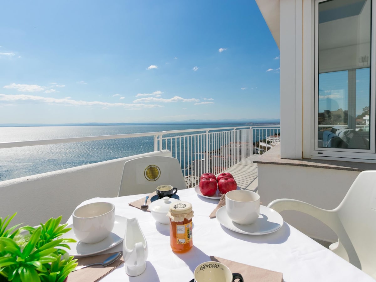 A dining table is set for breakfast on a sunny terrace, featuring white plates and bowls accompanied by apples and a jar of honey. The serene view of the sea is visible in the background under a clear blue sky, with gentle waves reflecting the sunlight.