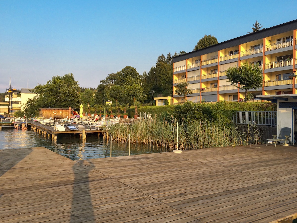 A view of the lake with a wooden pier extending into the water. Surrounding greenery and low shrubs are visible alongside the pier. In the background, a three-story building with balconies is situated, along with areas for sunbathing and relaxation.