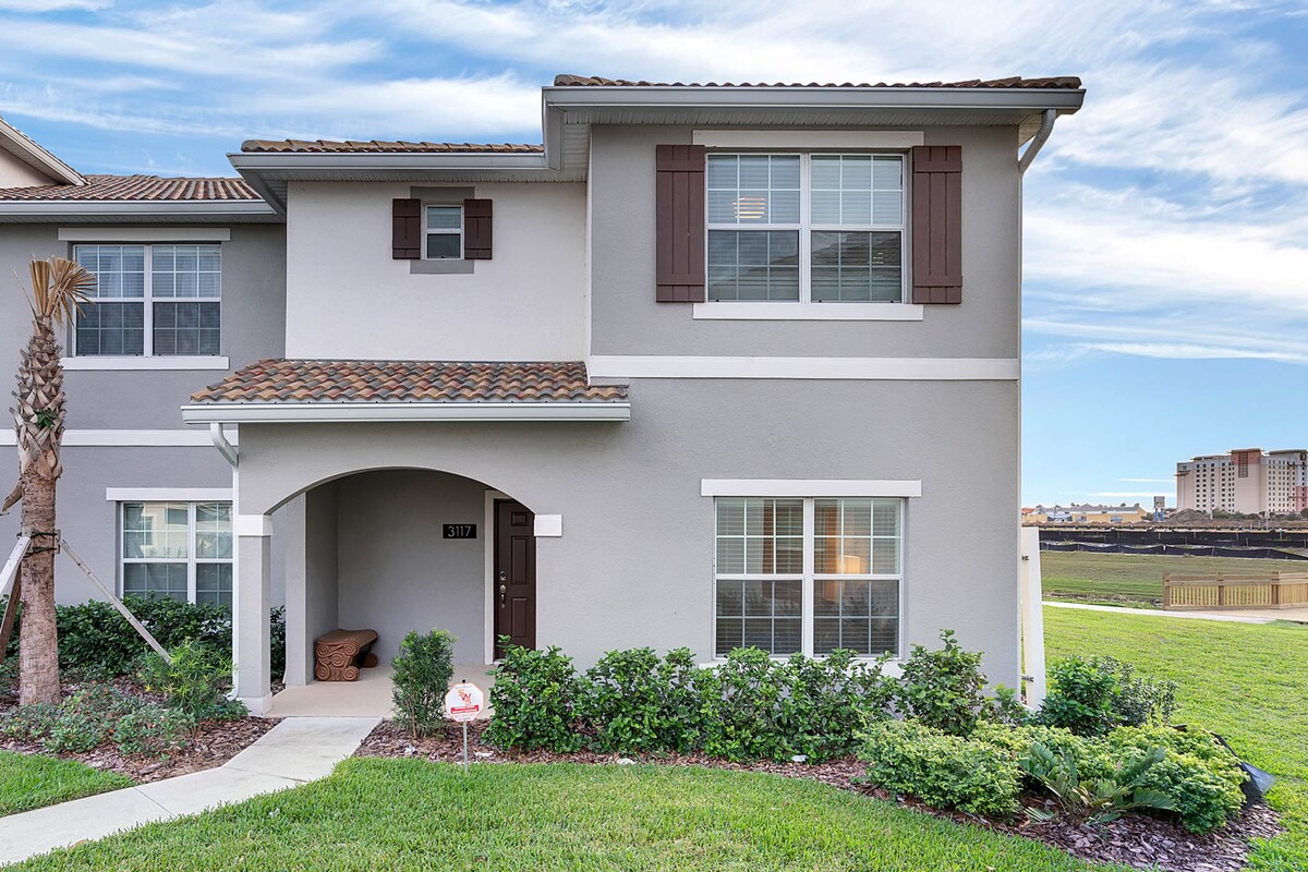 The exterior of the townhome is shown, featuring a covered entryway and well-maintained landscaping. The façade is painted in neutral tones, highlighted by white trim and brown shutters. A pathway leads up to the door, and a serene lake view is visible in the background.