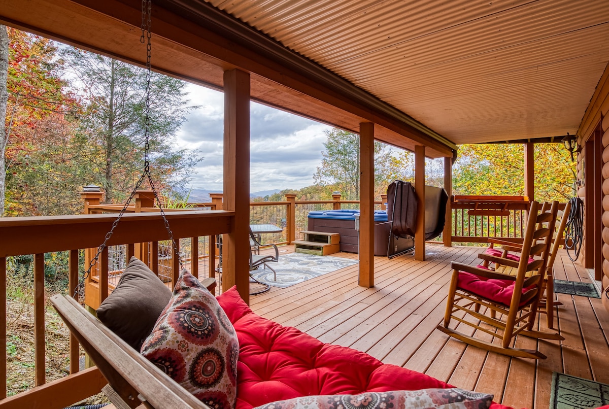 A covered porch features comfortable seating arrangements, including a swing and rocking chairs set against a backdrop of autumn-colored foliage. A hot tub is visible in the distance, accompanied by wooden railings that enhance the outdoor space's inviting atmosphere.