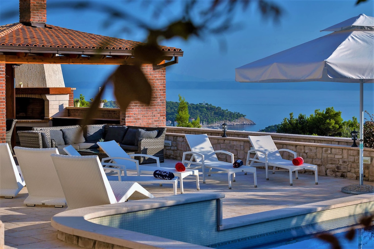 The outdoor terrace features a heated swimming pool and a water feature. Comfortable loungers and seating arrangements are positioned around the pool. A shaded area with a grill and summer kitchen offers additional dining space, surrounded by scenic views of the coast.