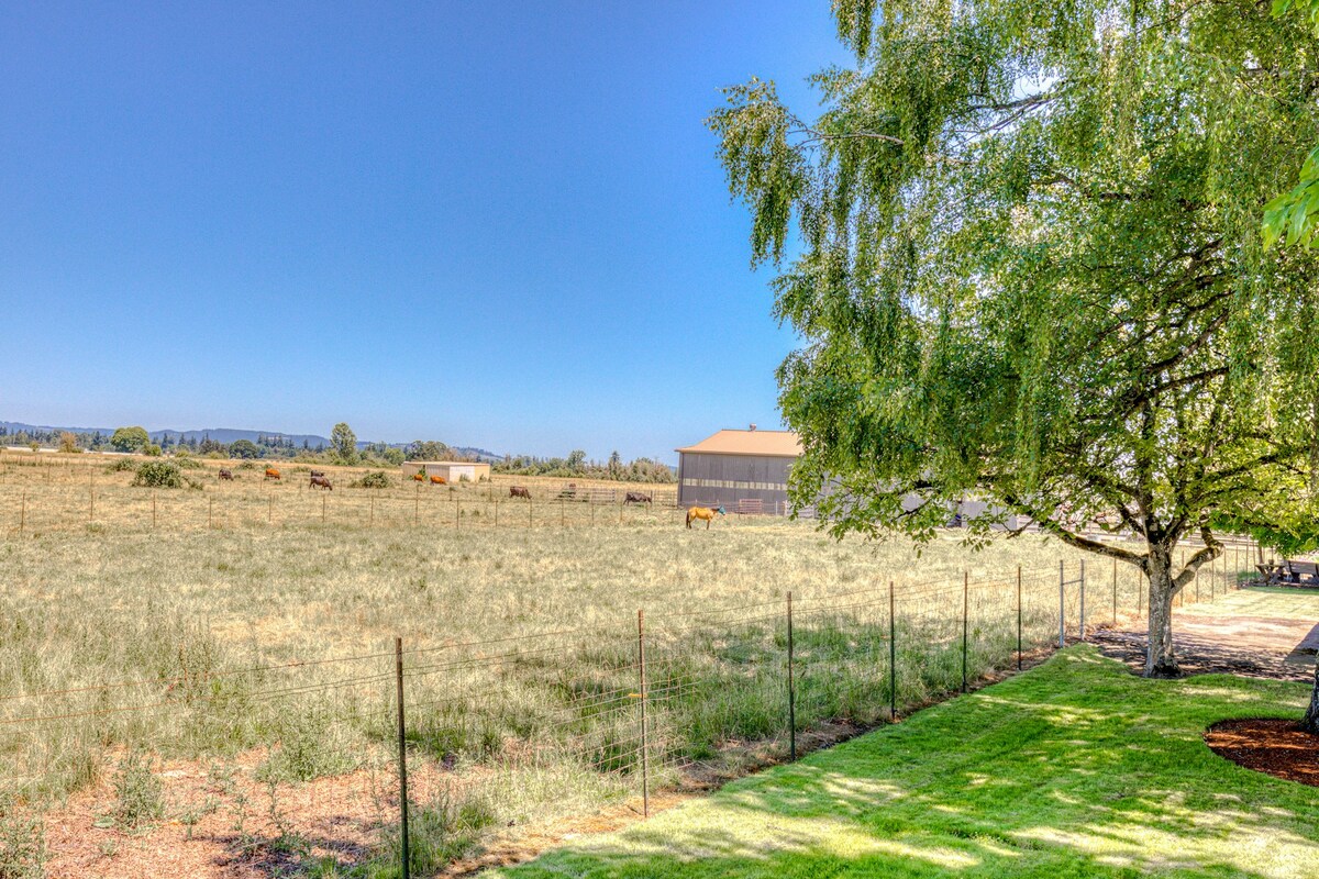 A serene view of the surrounding landscape showcases a sprawling open field with hints of grazing animals in the distance. A gentle breeze sways the lush grass near the tree, while a clear blue sky provides a calm backdrop to the rustic setting.