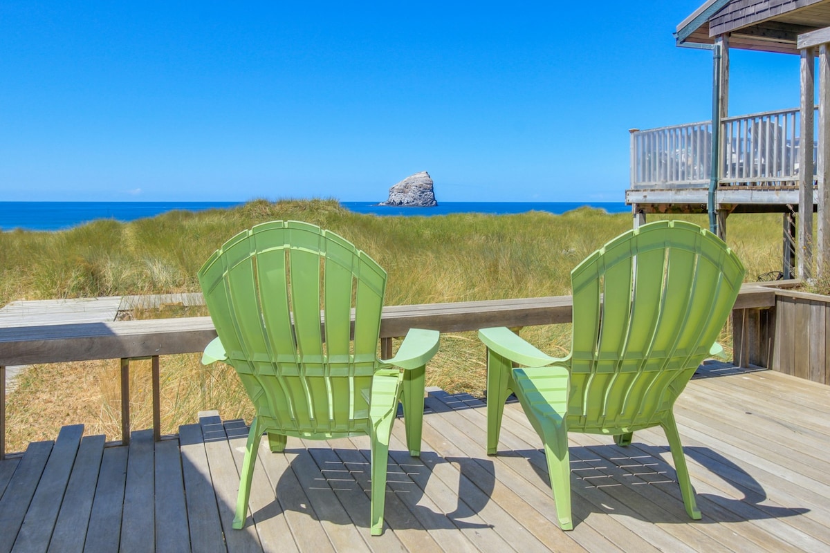 Two green Adirondack chairs are positioned on a wooden deck, facing a sandy beach and the ocean. Haystack Rock is visible in the distance against a clear blue sky, creating a serene coastal setting surrounded by tall grasses.