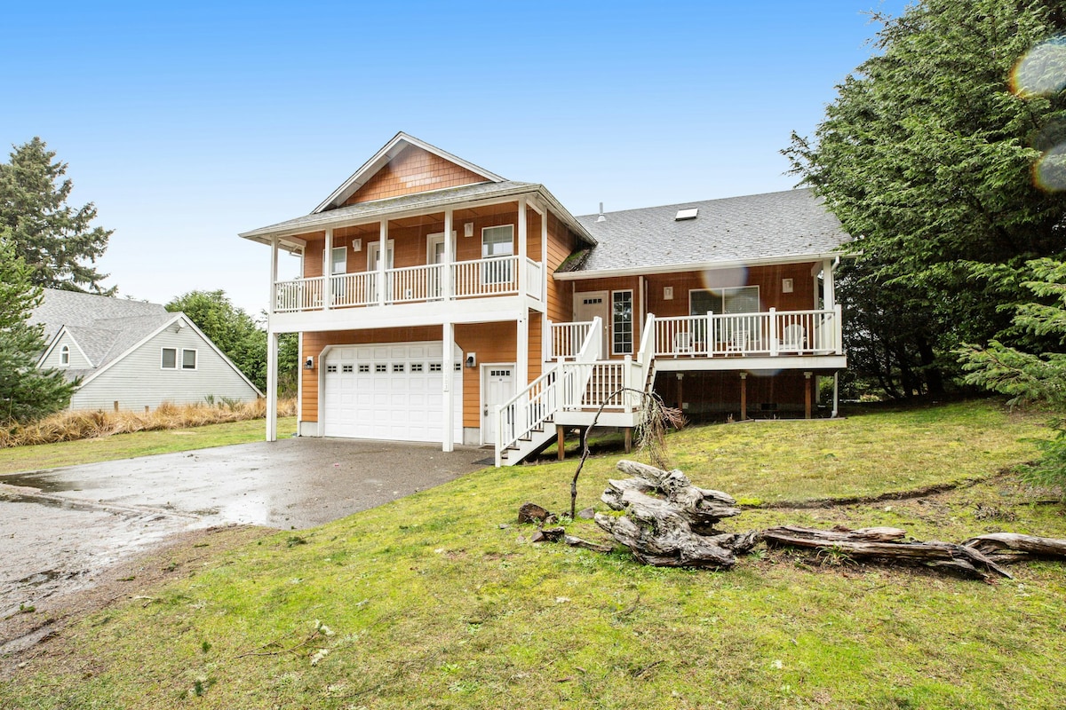 The exterior of a spacious two-story home is depicted, featuring a welcoming porch with white railings. A driveway leads to a two-car garage, surrounded by a grassy yard and tall trees, creating a serene, forested setting.