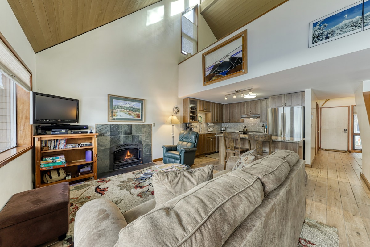 The living area features high vaulted ceilings and an inviting gas fireplace on a stone facade. A comfortable sectional sofa is positioned in front, while a TV and shelf with books are visible. Natural light enters through multiple windows, enhancing the open layout.
