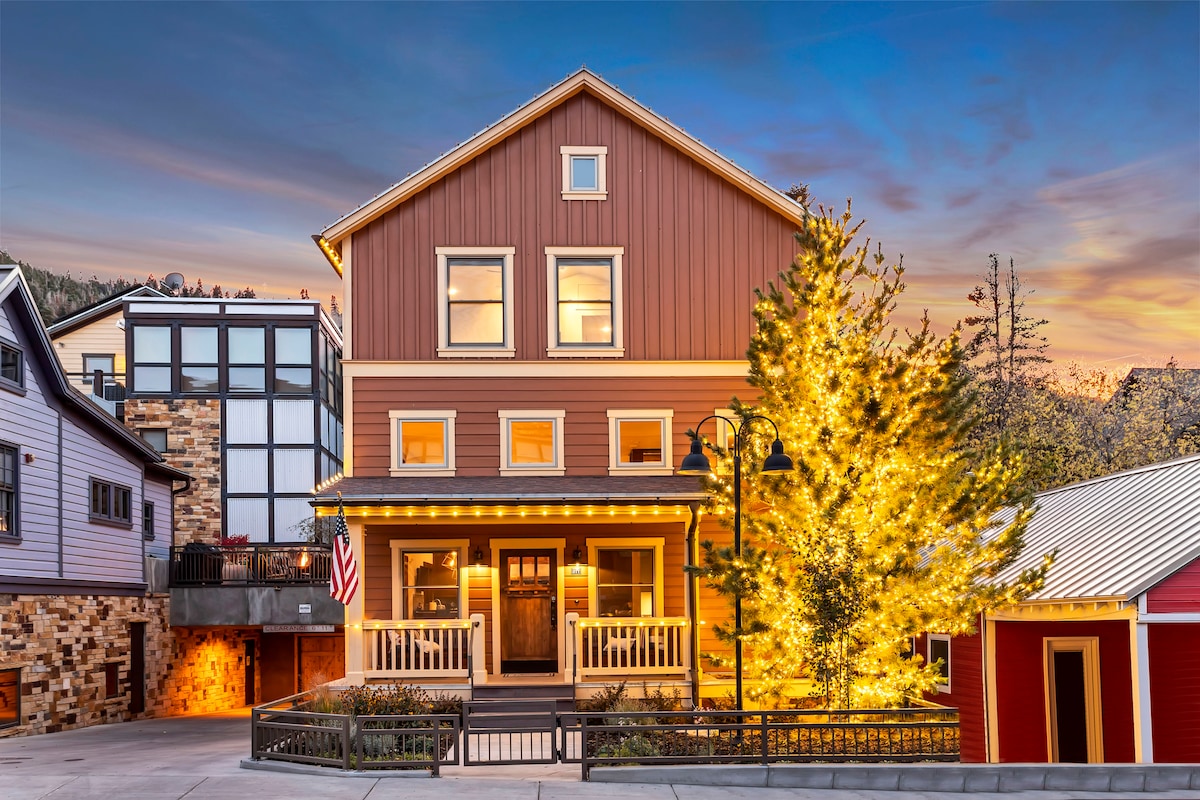 A charming three-story building features a mix of warm earth tones with large windows reflecting the sunset. Decorative outdoor lighting illuminates the front porch, complemented by an evergreen tree adorned with lights. Nearby structures showcase a blend of traditional and modern architecture.