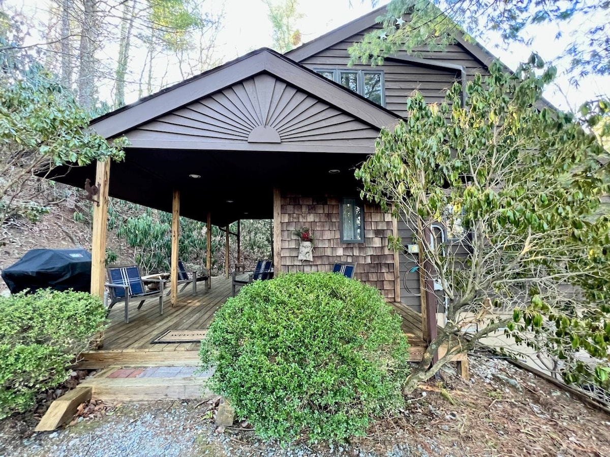 The entrance is framed by lush greenery, featuring a wooden porch with several blue chairs arranged for comfort. A black grill is positioned nearby, surrounded by well-maintained landscaping, providing a welcoming outdoor space.