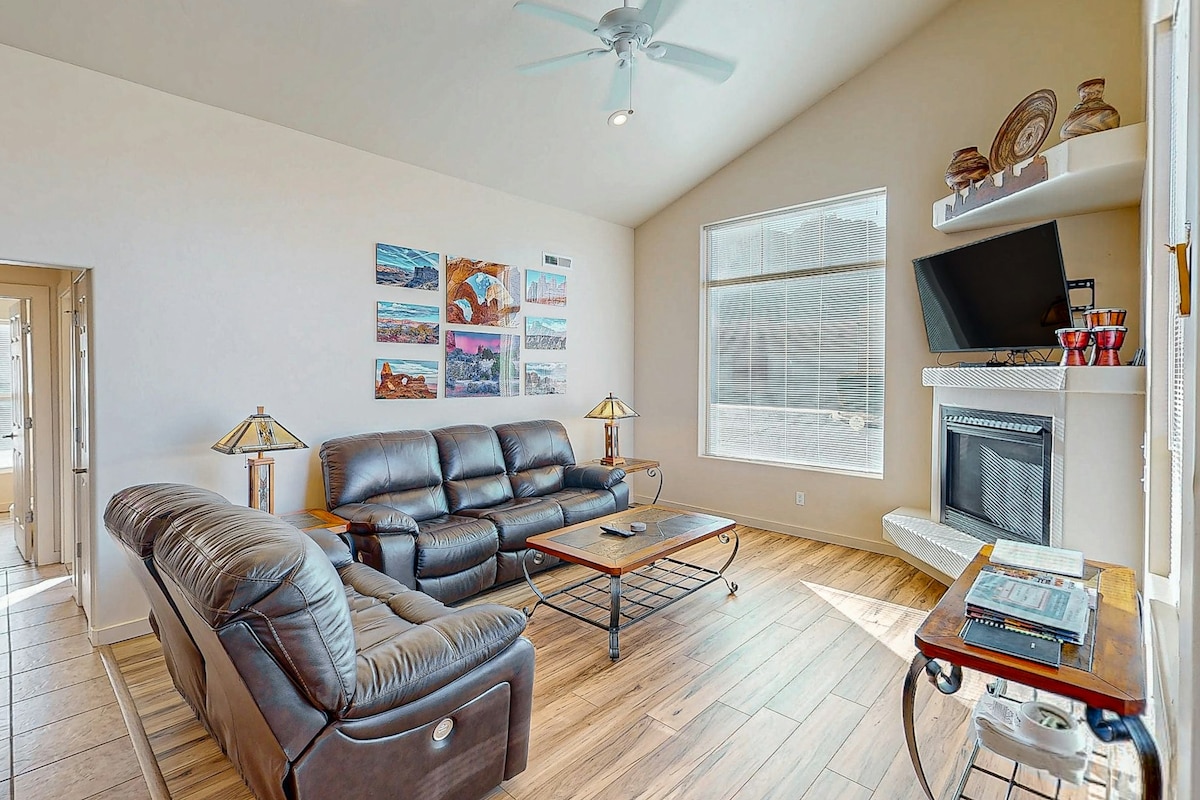 A bright living room is featured, showcasing comfortable leather seating arranged around a wooden coffee table. A cozy fireplace is positioned under a wall-mounted TV, while natural light fills the space through a large window. Decorative art pieces add subtle color to the walls.