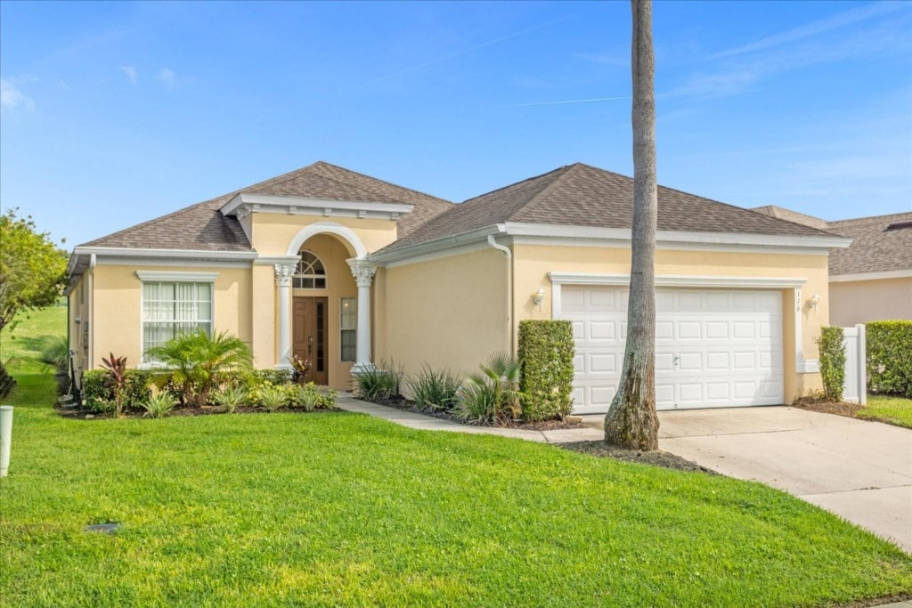 A single-story villa is showcased, featuring a light-colored facade and a well-manicured lawn. Lush greenery lines the pathway leading to the front door, while a spacious driveway provides ample parking. A palm tree adds a tropical touch to the entrance.