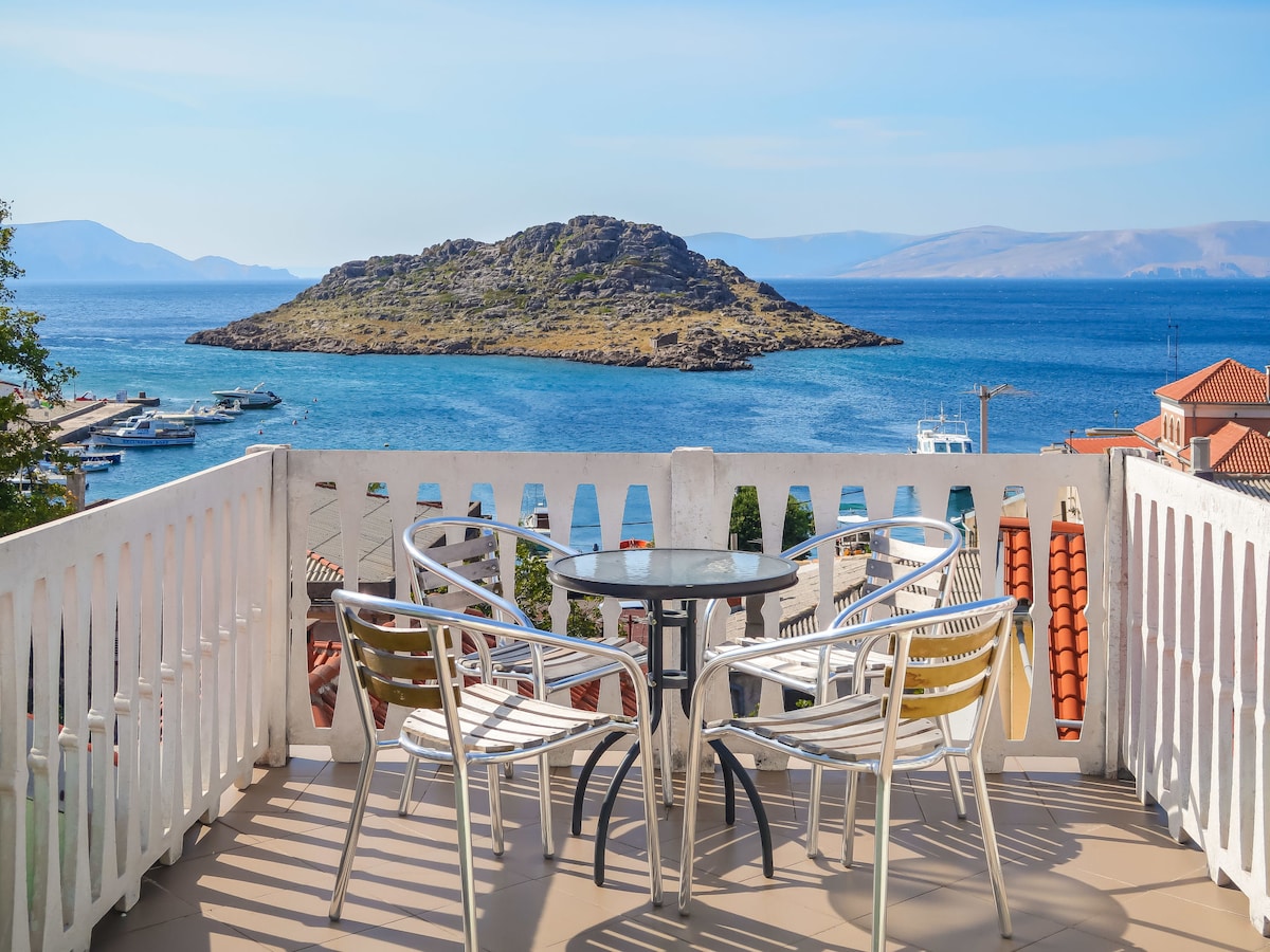 A sunlit terrace features a glass-top table surrounded by four metal chairs. The view includes a serene sea and a small island in the distance, complemented by the blue sky and distant mountains.