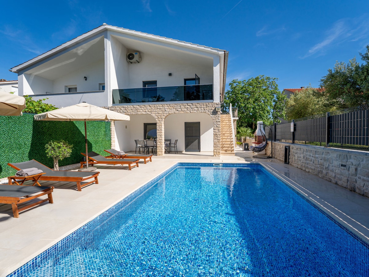A pool area is presented with a heated swimming pool surrounded by loungers and parasols. The semi-detached house, featuring a balcony, is visible against a clear blue sky. Greenery borders the space, providing a tranquil setting.