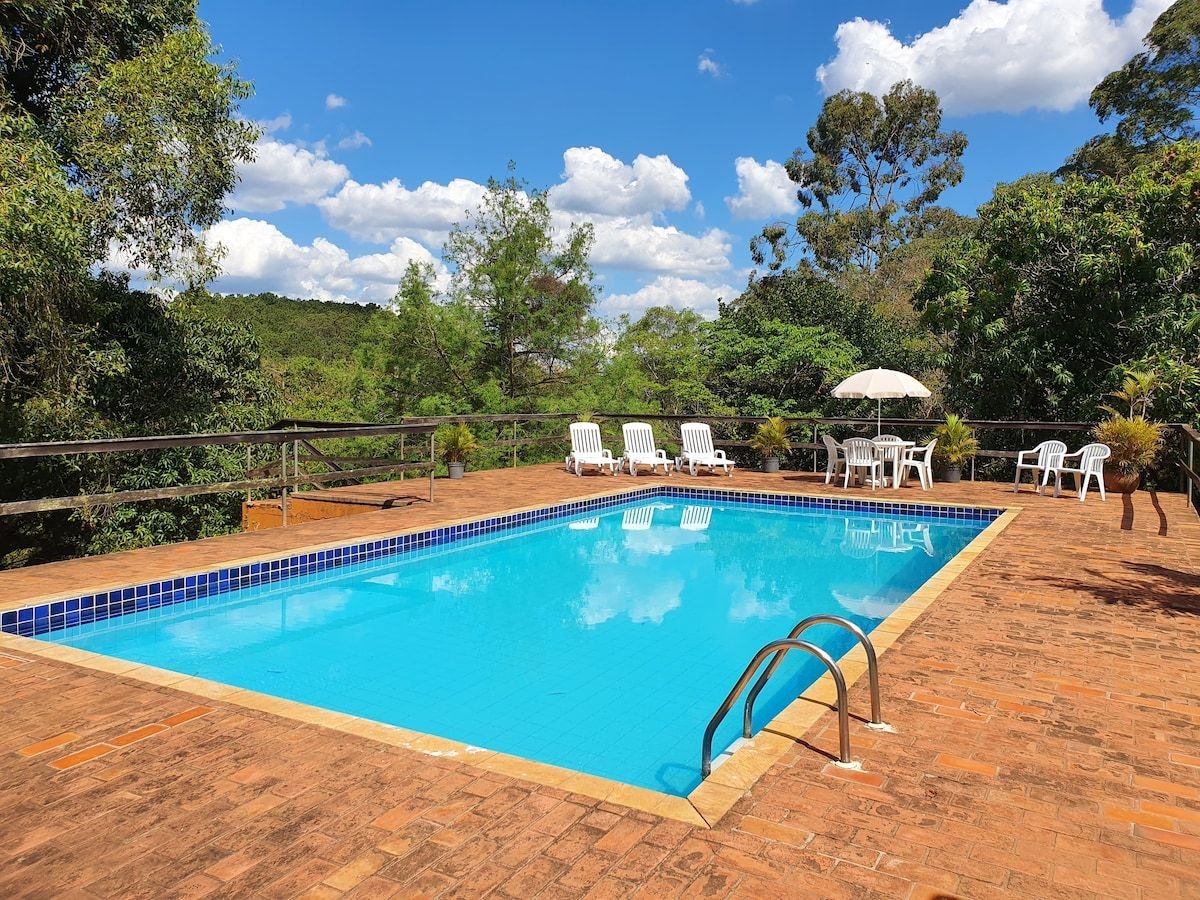 A spacious outdoor swimming pool is surrounded by a wooden deck, featuring light blue tiles. Several lounge chairs are positioned on one side, with a table and chairs under an umbrella nearby. Lush greenery and trees frame the peaceful setting.