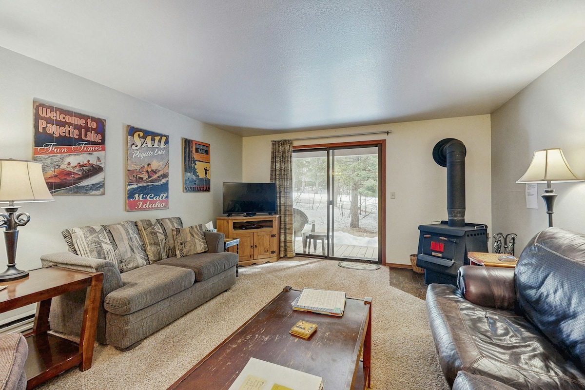 A living room area features a comfortable couch and armchair, with a wood-burning stove visible in the corner. A coffee table and a TV stand organized with entertainment options are present. Natural light enters through sliding glass doors, leading to an outdoor deck.