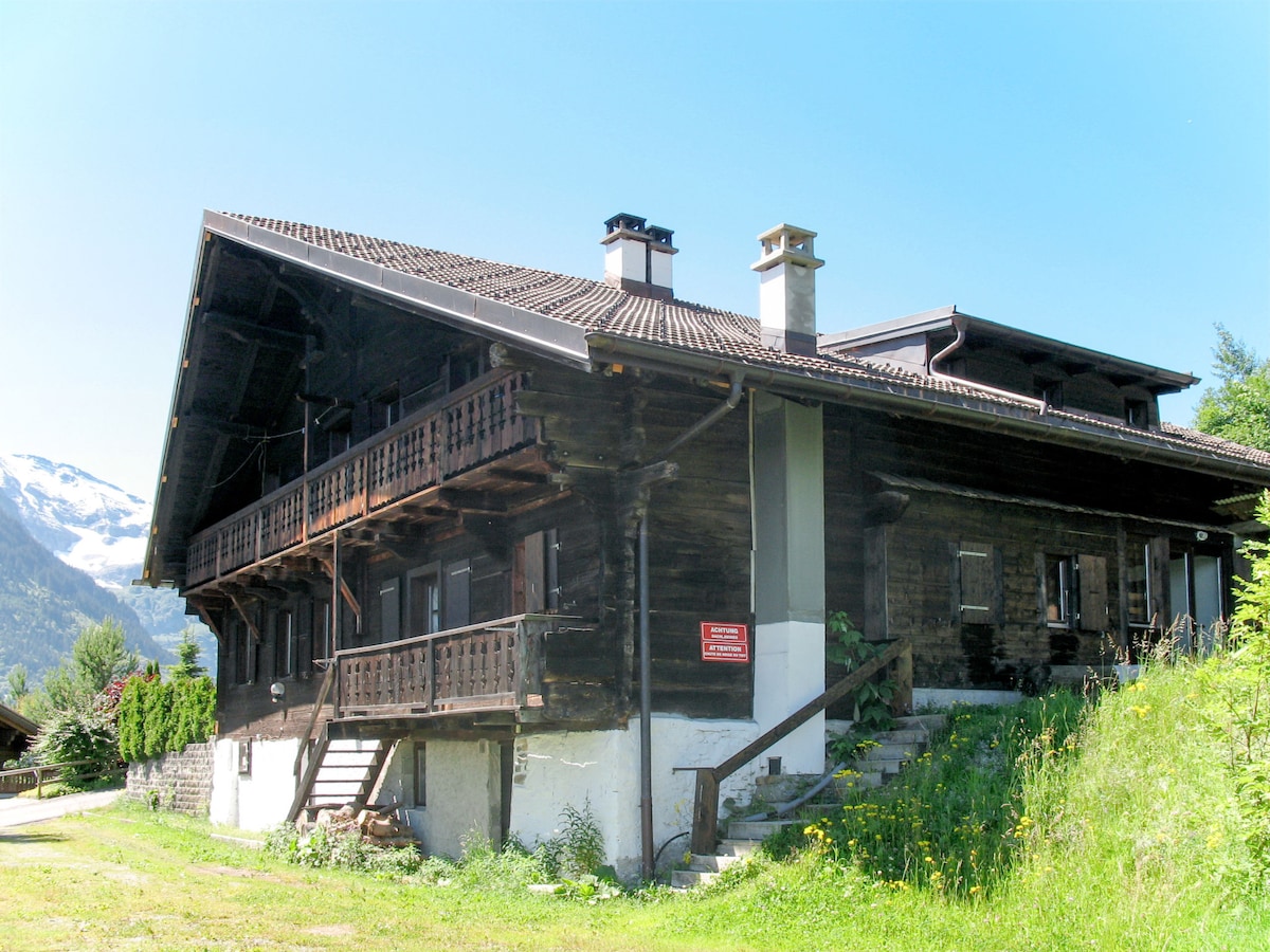 A large, rustic chalet exudes charm with its wooden exterior and expansive balconies. The structure is built on two levels, featuring multiple entrances and a sloped path leading to the front entrance. Surrounded by greenery, it offers a scenic view of the mountains in the background.