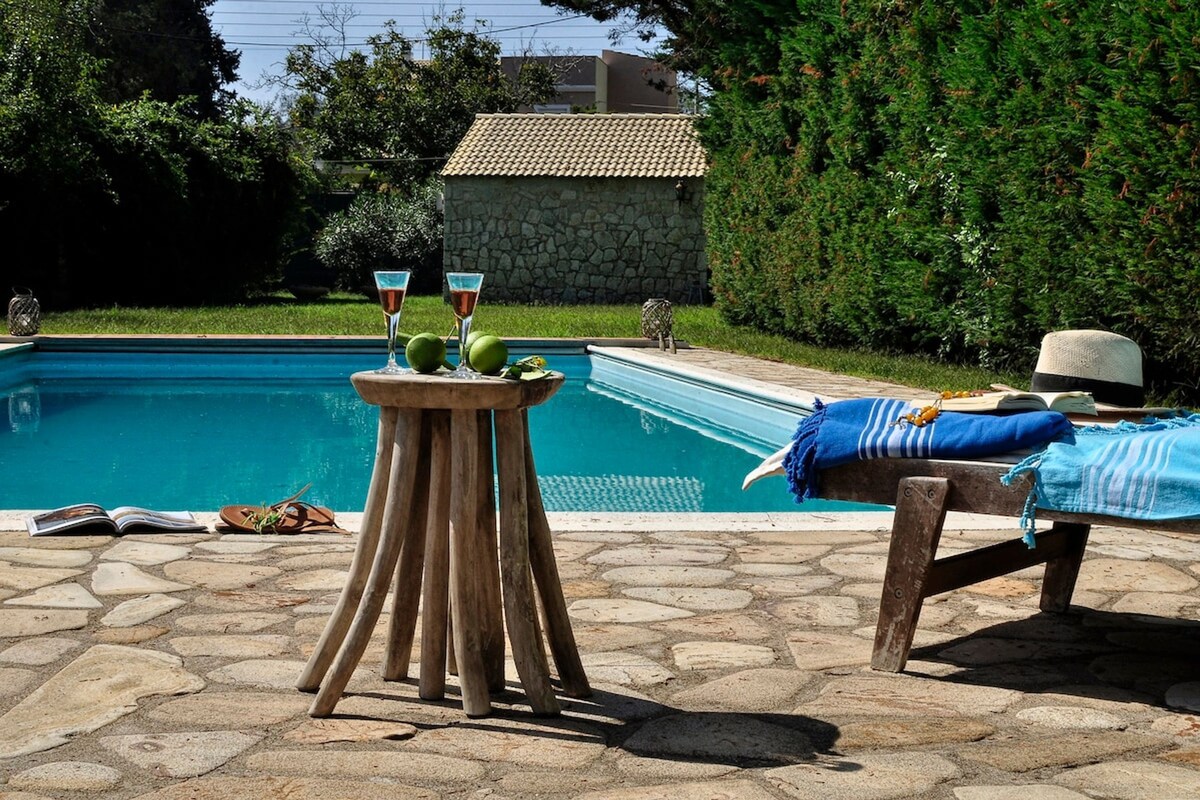 A tranquil poolside area is featured, with a refreshing blue pool surrounded by well-maintained greenery. A rustic wooden table holds two drinks and fresh limes. A sunbed, adorned with a blue striped towel and a straw hat, invites relaxation under the sun.