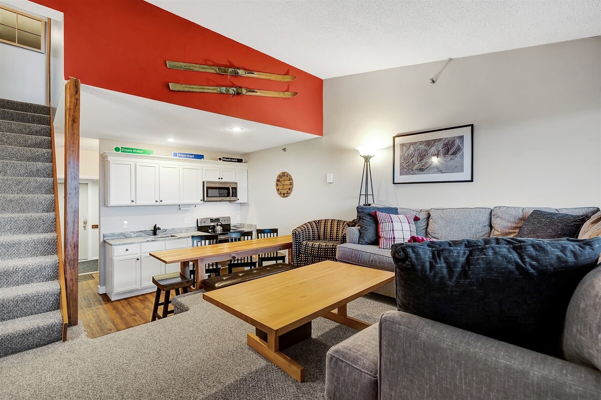 A spacious living area features a large L-shaped sectional sofa complemented by a coffee table. A dining table with multiple seats is positioned nearby, while a well-equipped kitchen area can be seen in the background. Natural light streams in from a nearby source, enhancing the cozy ambiance.