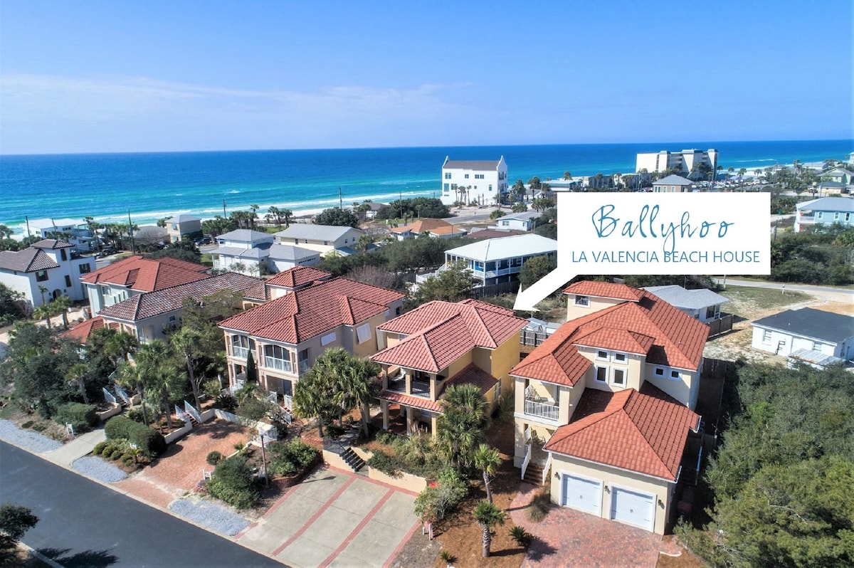 An aerial view showcases the vibrant roofs of the Mediterranean-style villas within the gated community of La Valencia. The turquoise Gulf of Mexico sparkles in the background, framed by lush greenery and sandy beaches, positioning Ballyhoo Villa conveniently near the shore.