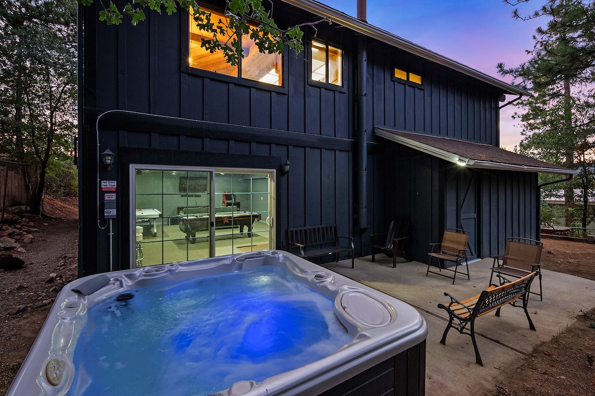 An outdoor space features a relaxing hot tub with illuminated water, surrounded by a concrete patio. Nearby, two benches provide seating options. Large windows on the upper level offer glimpses of warm indoor lighting, suggesting a cozy ambiance within the cabin.