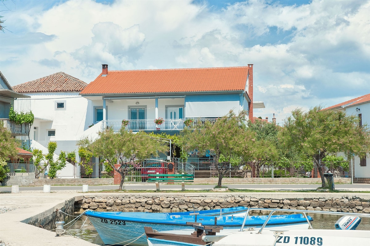 A two-story building is depicted, featuring a balcony adorned with potted plants. The exterior is painted in light blue, contrasted by a red-tiled roof. A paved pathway leads to the water, where two boats are moored along the stone wall.