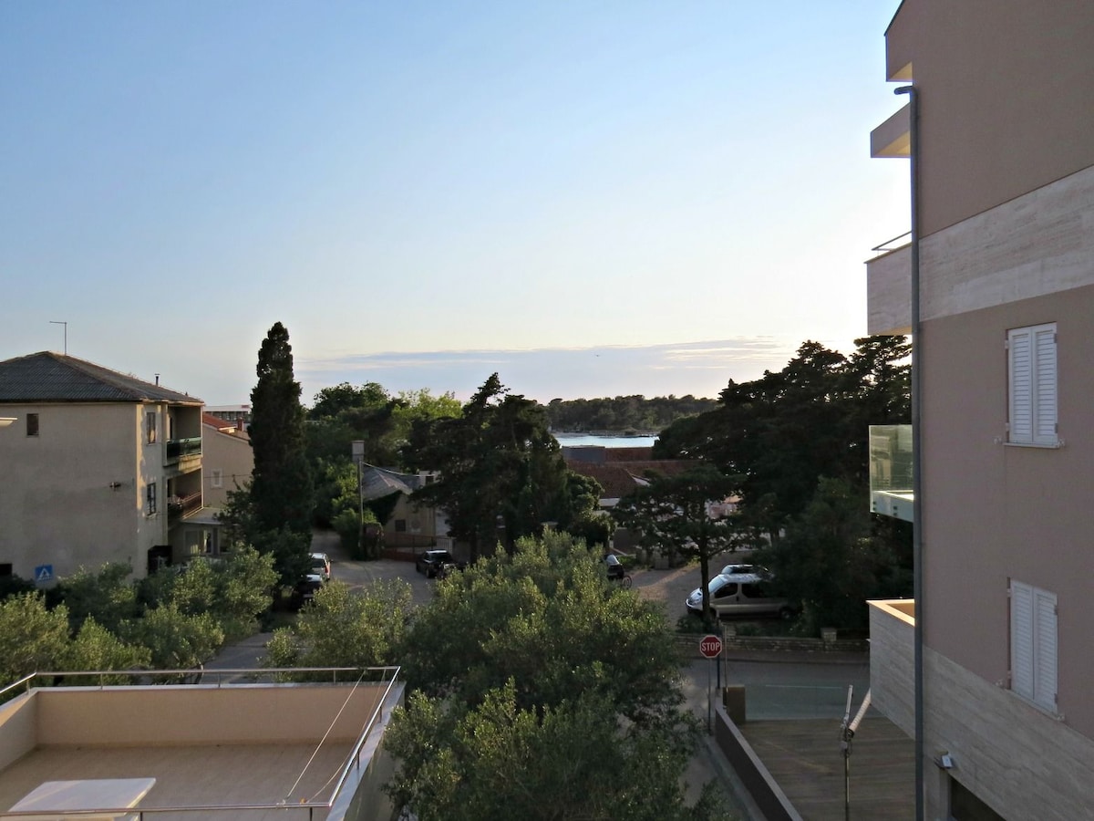 An outdoor view showcases a peaceful neighborhood with lush greenery and residential buildings. The horizon reveals a glimpse of the sea, framed by trees, while a gentle light illuminates the scene, suggesting early morning or late afternoon.