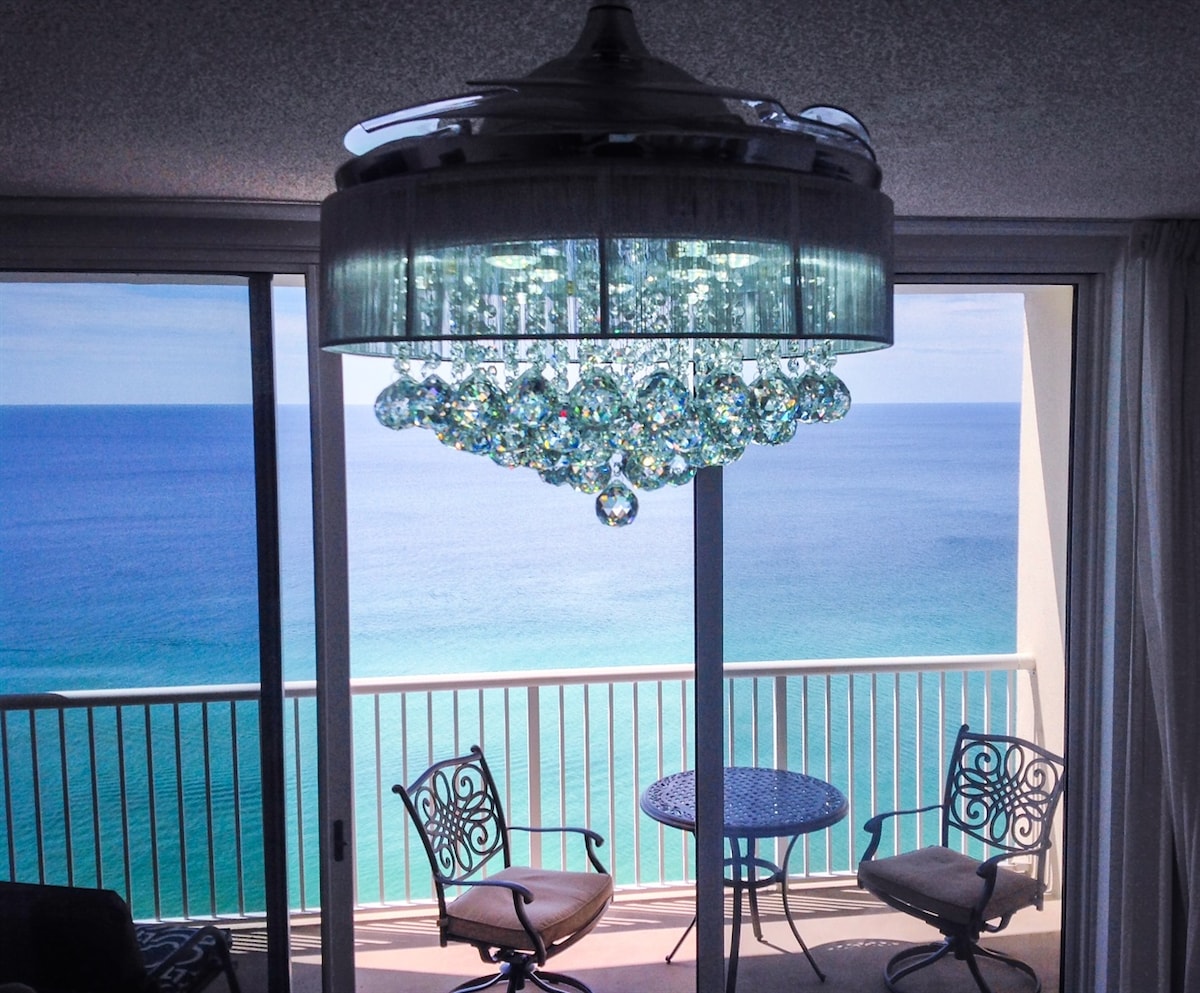A modern chandelier adorned with crystal accents hangs from the ceiling, illuminating the dining area. A circular blue table with ornate chairs overlooks the expansive blue waters of the Gulf through large glass doors, enhancing the beachfront ambiance.