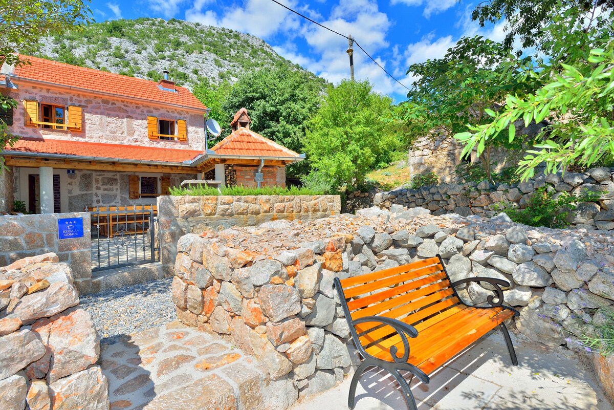 A stone house is framed by a lush green landscape, featuring a welcoming wooden bench positioned in a stone-paved area. The house's rustic architecture is complemented by orange roof tiles and wooden shutters, providing a harmonious blend with the surrounding nature.