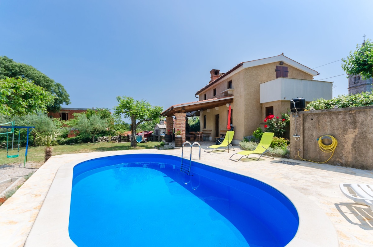An outdoor pool is surrounded by a sunny terrace, featuring two lounge chairs in a vibrant green hue. The house's exterior showcases rustic walls with floral accents and shaded seating areas. Lush greenery and garden elements create a relaxing atmosphere in the background.