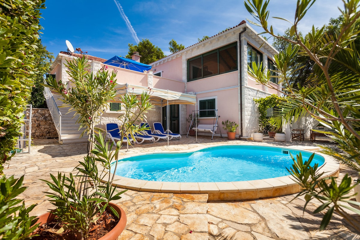 A private outdoor area features a circular pool surrounded by stone pathways and lush greenery. Sun loungers are positioned nearby under an awning, providing a shaded spot for relaxation. The villa's exterior is painted in soft pink, harmonizing with the vibrant surroundings.
