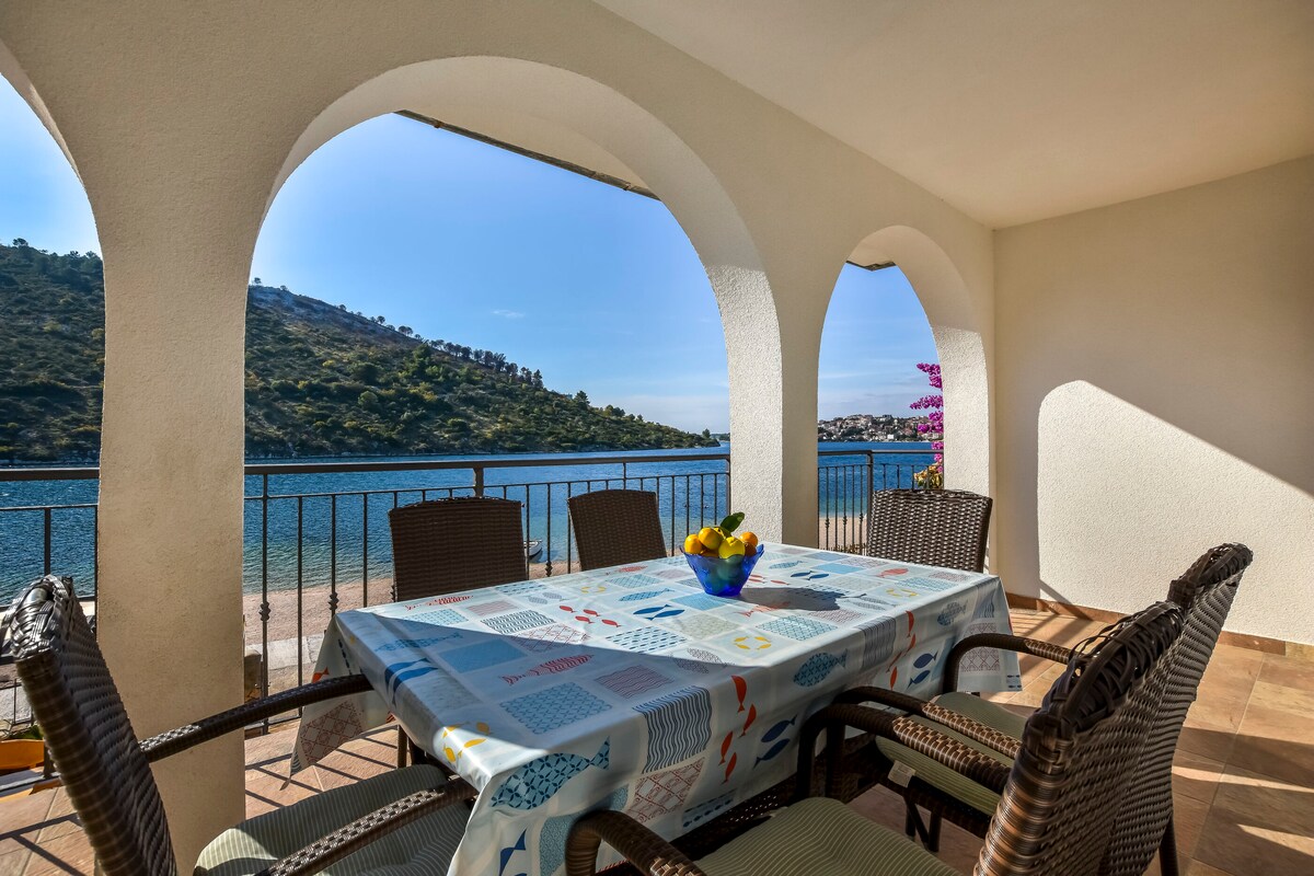 An outdoor terrace is displayed, featuring a table set with a colorful tablecloth. Surrounding the table are woven chairs. A view of calm waters and lush hills is visible through arched openings, enhancing the inviting atmosphere of the space.