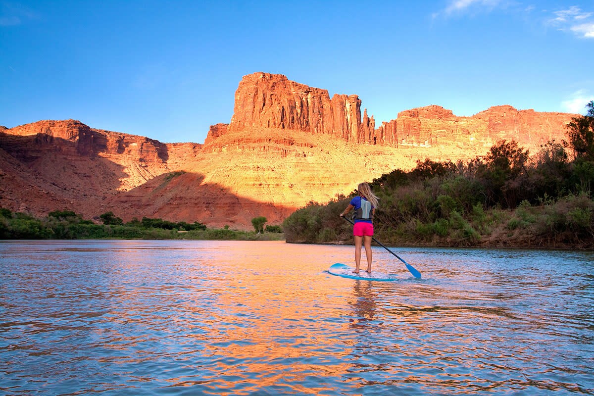 A paddleboarder glides across calm waters, surrounded by towering red rock formations bathed in warm evening light. The landscape features mountainous terrain with vibrant colors reflecting on the water's surface, creating a serene outdoor setting.