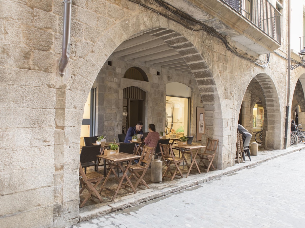 A charming outdoor dining area is depicted with wooden folding tables and chairs arranged beneath a stone archway. The rustic architecture features original stone walls, and patrons can be seen enjoying their meals. Soft lighting enhances the inviting atmosphere of this historic setting.