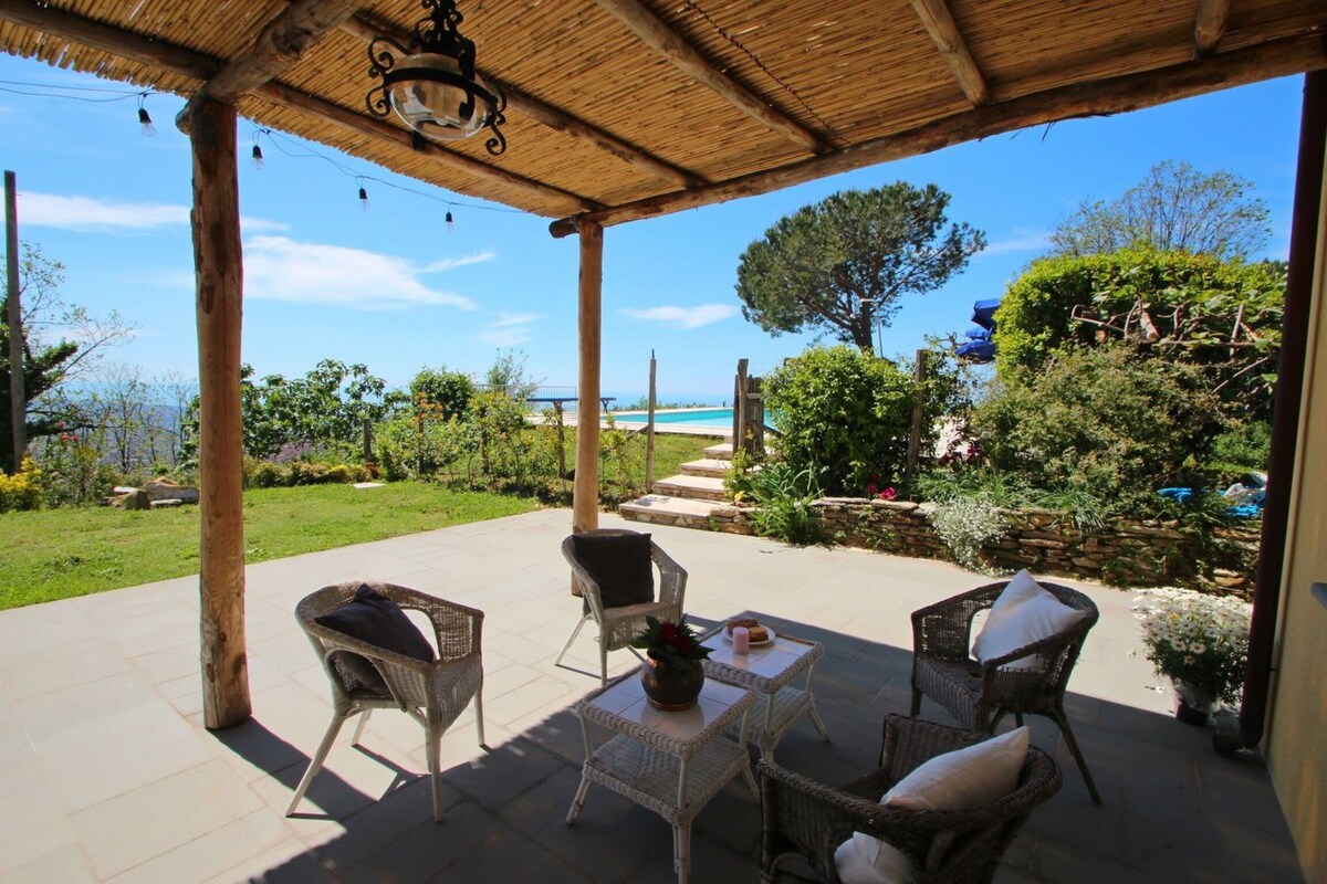 A covered porch area features a set of four wicker chairs around a low table, offering a comfortable space for relaxation. A sweeping view of the greenery and distant sea is visible through the open porch, complemented by clear blue skies.