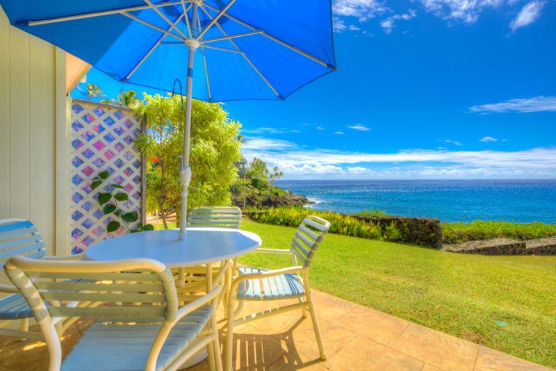 An outdoor seating area features a round table accompanied by four light blue chairs. A large blue umbrella provides shade, while a lush green lawn leads to the ocean, visible in the background under a clear sky dotted with a few clouds.