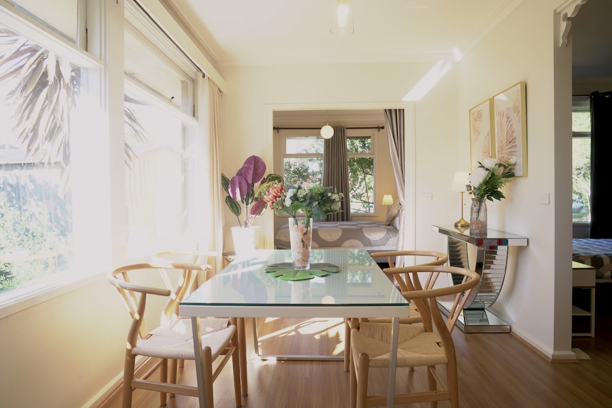 A bright dining area features a glass table surrounded by light wooden chairs. Natural light enters through large windows adorned with soft curtains. A decorative vase with greenery sits at the center of the table, emphasizing the room's inviting ambiance.