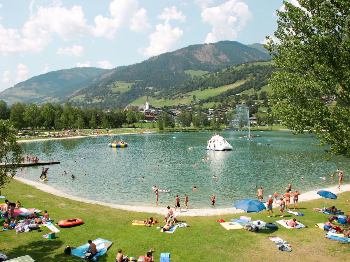 A scenic lakeside view features a clear water area surrounded by a grassy beach. Visitors enjoy the warmth of the sun while lounging on colorful blankets. A floating play structure and various devices for water activities are visible, with mountains providing a backdrop of lush greenery.