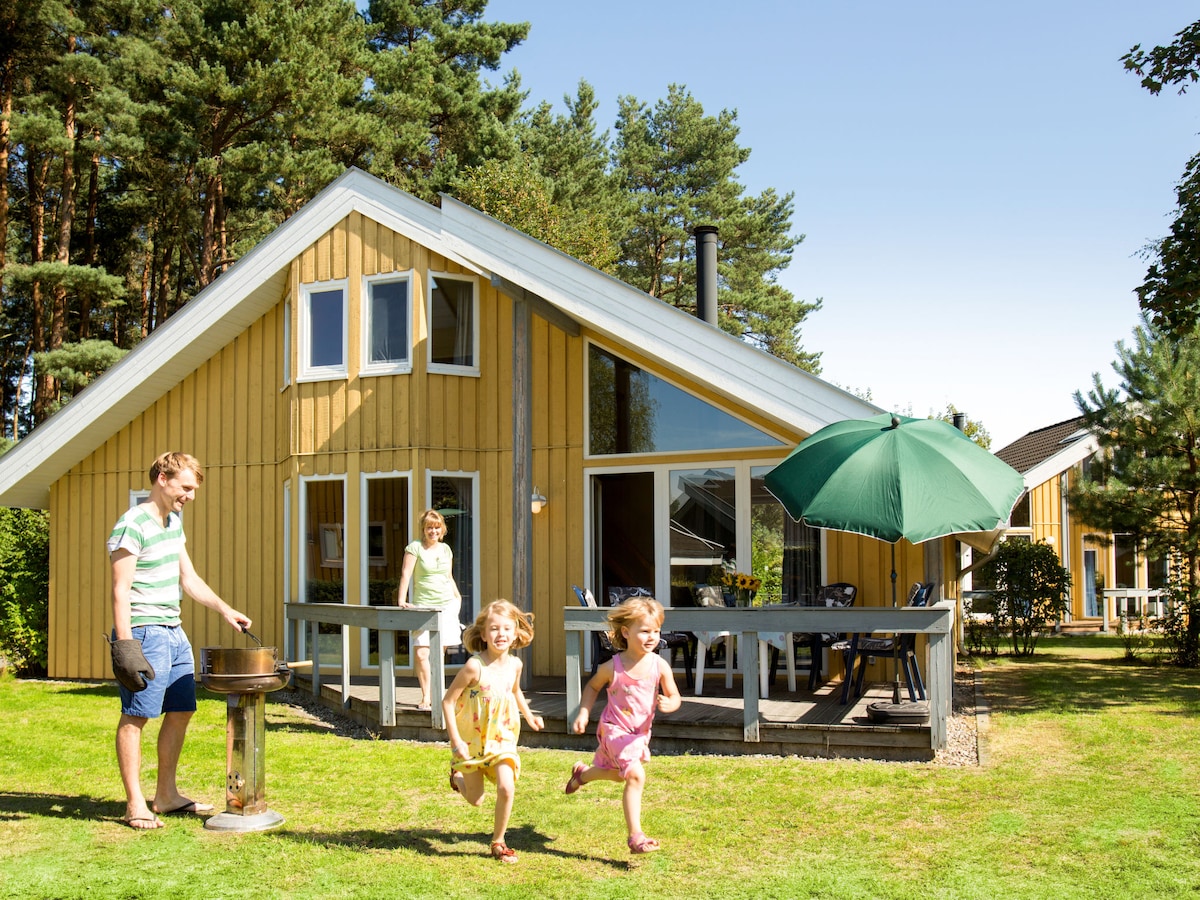 A charming semi-detached house with a yellow wooden façade sits surrounded by trees. A spacious deck features outdoor furniture and a large umbrella. Children are seen playing on the lawn, while a barbecue grill is positioned nearby, inviting family gatherings.