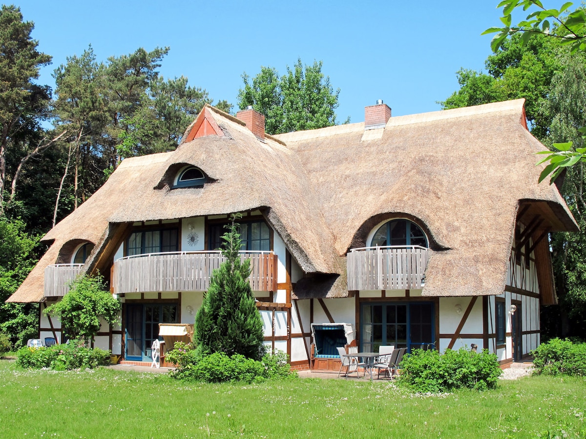 The exterior of a charming two-story building features a distinctive thatched roof and framed in light-colored wood. Two balconies extend from the upper level, overlooking a well-maintained lawn with greenery. Tall trees surround the property, providing a natural backdrop.