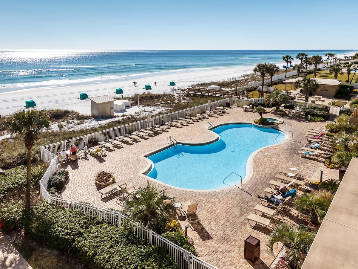 An inviting outdoor pool is surrounded by paver stones and tropical landscaping. Beach chairs and umbrellas are set up along the shoreline, with the Gulf of Mexico visible in the background under clear skies. Palm trees provide a lush backdrop to the serene setting.