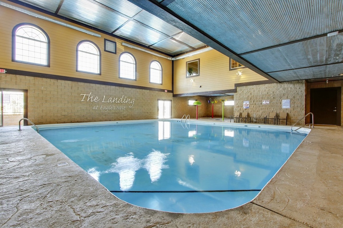 An indoor heated saltwater pool is depicted, featuring a smooth lightly textured surface. Large windows along the upper wall allow ample light to fill the space. The pool area is furnished with a few seating options nearby, enhancing the inviting ambiance for relaxation and leisure.
