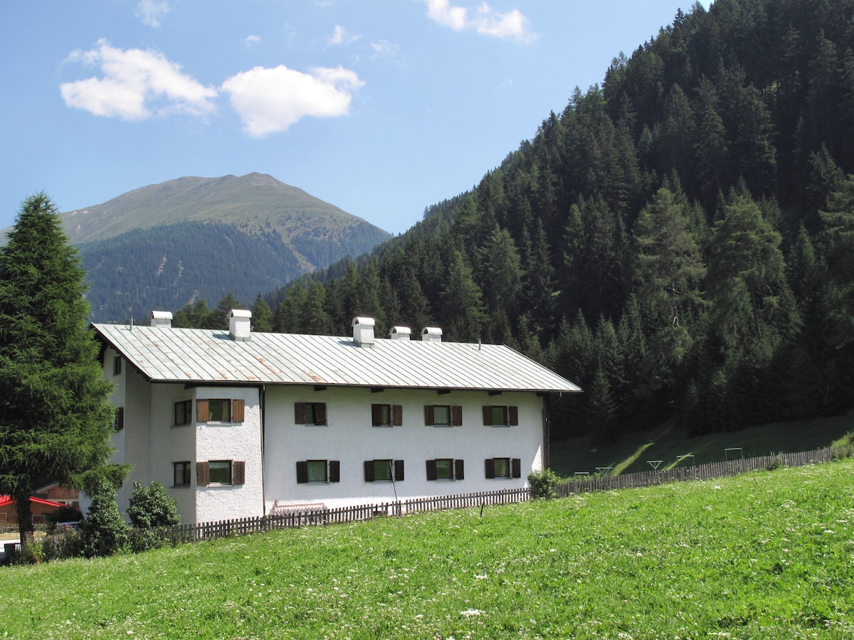 A multi-story building with a light-colored exterior is situated amidst a green field. Windows with green shutters are visible, along with a metal roof. The backdrop features a mountainous landscape dotted with trees, enhancing the serene environment.