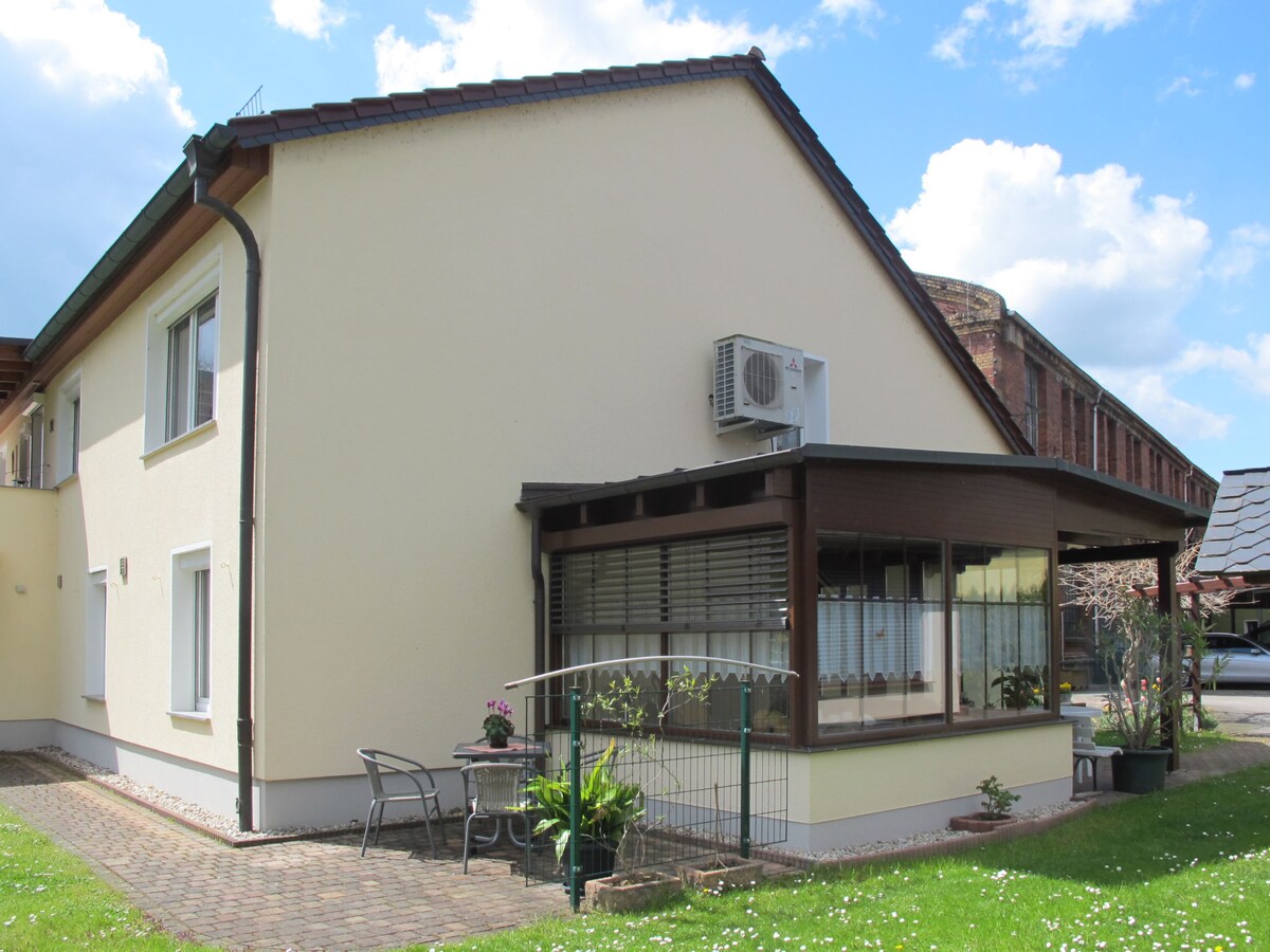 The exterior of the two-family house features a light-colored facade with a sloped roof and windows. A winter garden is visible, with chairs placed outside on a brick patio surrounded by greenery. An air conditioning unit is mounted on the wall.