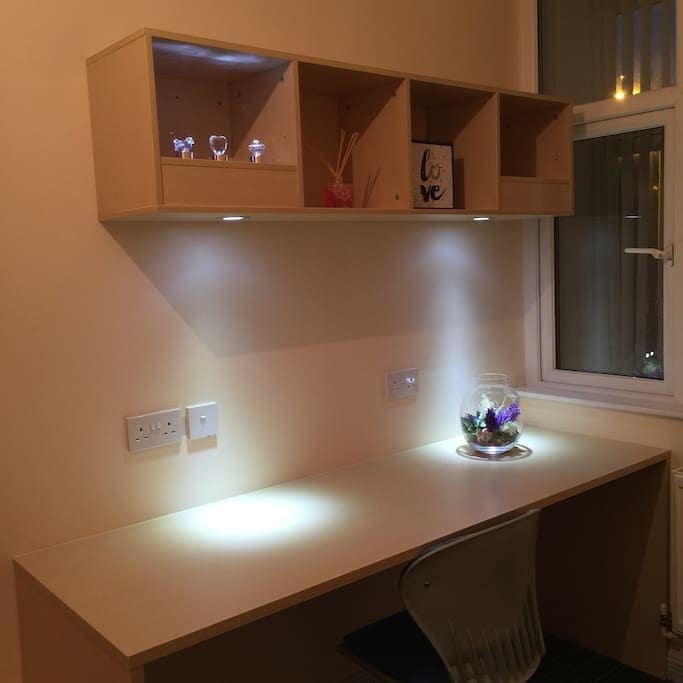 A minimalist workspace is presented, featuring a clean white desk with a modern clear chair. A shelf above holds decorative items and a small arrangement of plants, softly illuminated along with the desk's integrated lighting, creating a functional and inviting area for work.